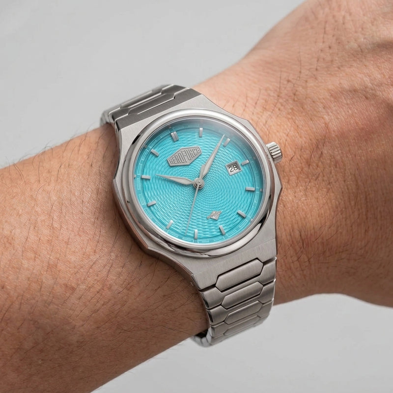 A silver stainless steel wristwatch with a textured turquoise blue dial, shown worn on a person's wrist against a neutral light gray background.