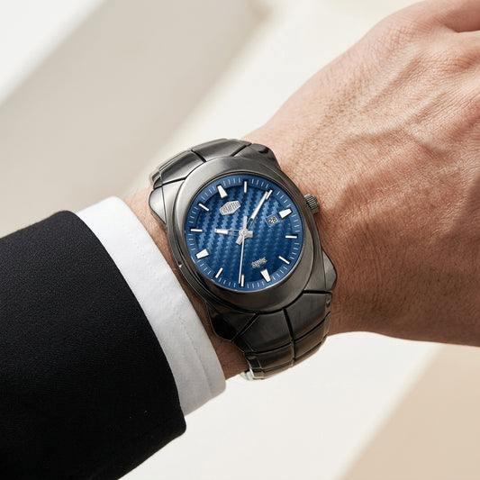 A blue watch with a textured dial and integrated bracelet worn on a person's wrist, shown against a softly blurred light background. The person is dressed in a black suit jacket and white dress shirt.