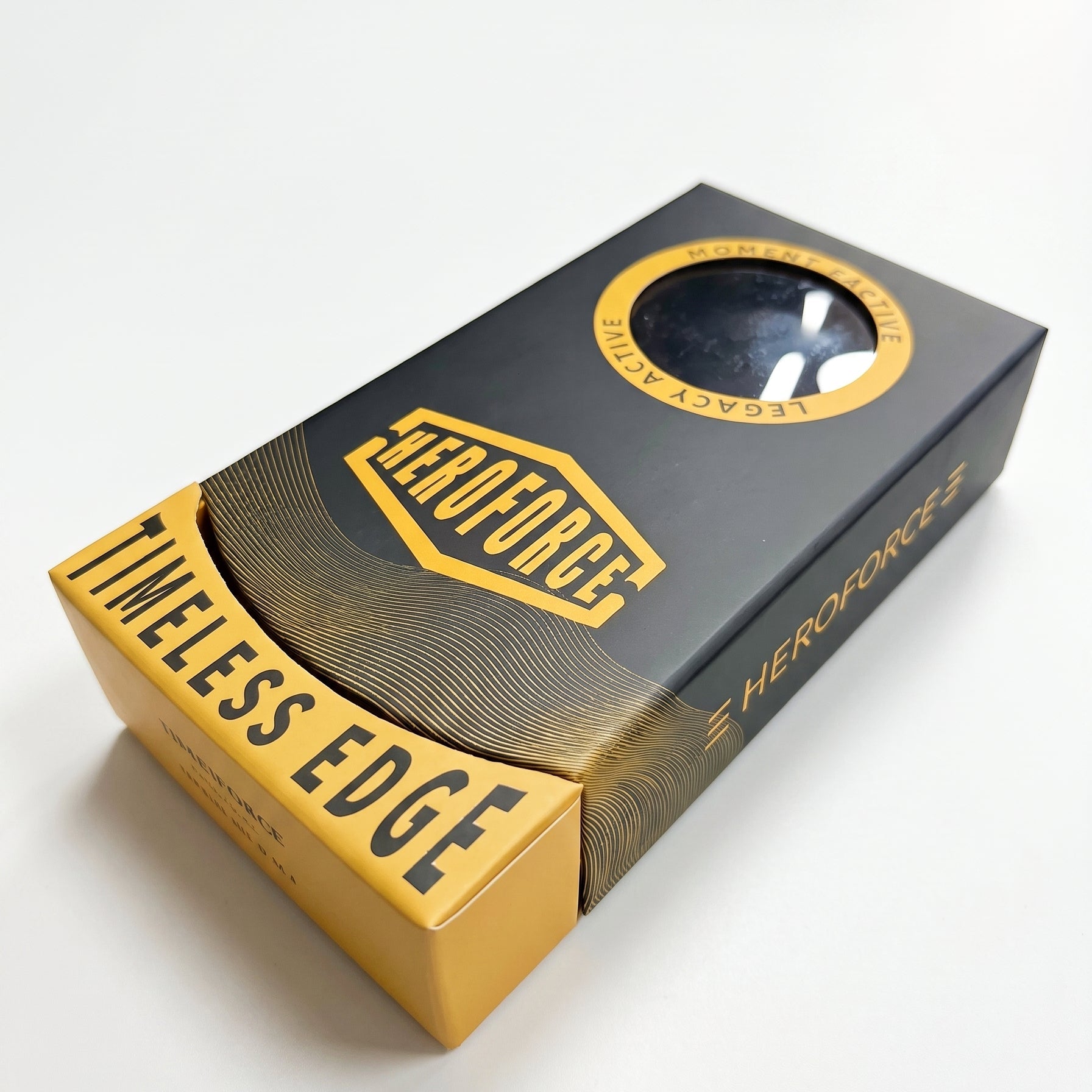 A boxed product labeled HEROFORCE and TIMELESS EDGE, shown at an angle on a clean white background. The box is primarily black with prominent gold/yellow panels and text, plus a circular window near the top.