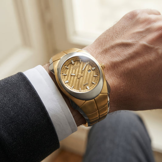 A close-up of a man's wrist wearing a luxury two-tone watch with a gold textured dial, gold and silver-tone case, and gold-tone link bracelet, styled with a white dress shirt cuff and dark suit jacket.