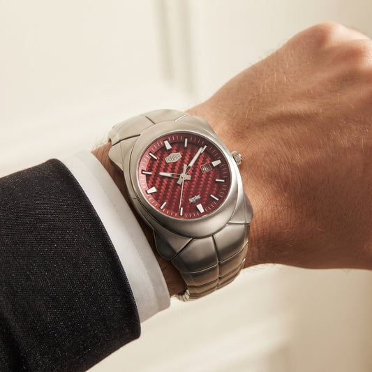 A close-up of a man's wrist wearing a silver-toned metal watch with a textured red dial and silver bezel, styled with a white dress shirt cuff and black suit jacket.