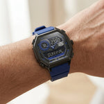 A close-up of a rugged digital watch with a blue strap and black case, worn on a person's wrist against a softly blurred light background.