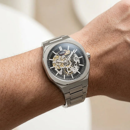 A close-up of a wrist wearing a silver-toned skeleton watch with a black dial and visible gold-tone mechanical gears.