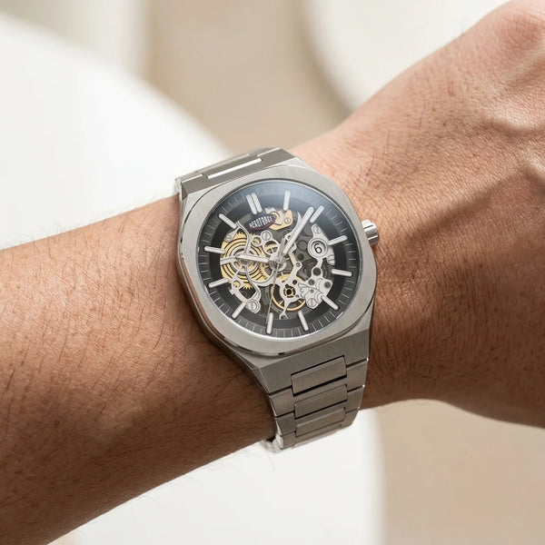 A close-up of a wrist wearing a silver-toned skeleton watch with a black dial and visible gold-tone mechanical gears.