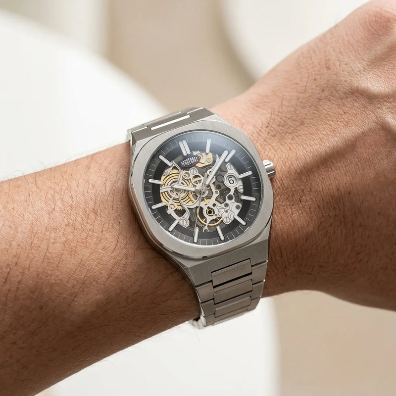A close-up of a wrist wearing a silver-toned skeleton watch with a black dial and visible gold-tone mechanical gears.