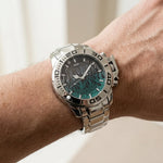 A close-up of a wrist wearing a stainless steel analog watch with a dark dial and a teal-green wave pattern, set against a soft, out-of-focus light background.