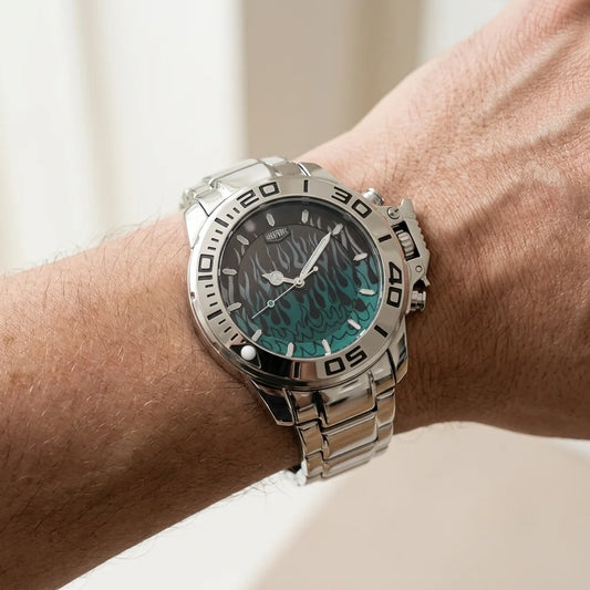 A close-up of a wrist wearing a stainless steel analog watch with a dark dial and a teal-green wave pattern, set against a soft, out-of-focus light background.