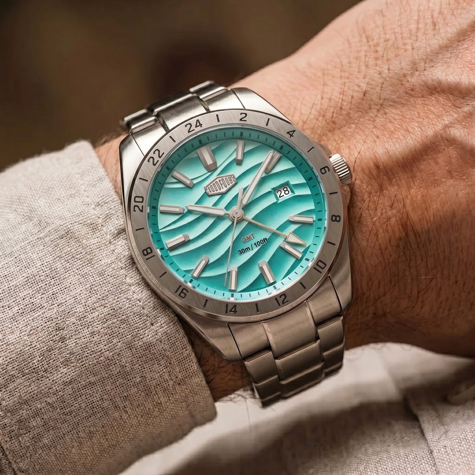 A close-up of a wrist wearing a stainless steel watch with a bright turquoise dial and matching metal bracelet, shown against a soft beige shirt and blurred background.