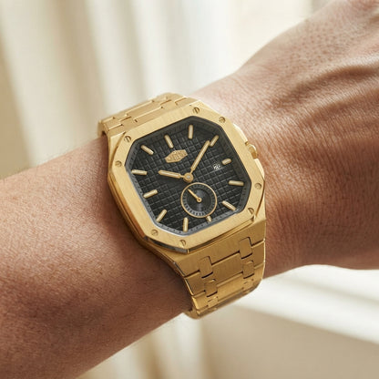 A gold-tone stainless steel watch with a textured black dial and matching gold link bracelet, shown worn on a person's wrist against a soft beige background.