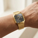 A gold-tone watch with a square black dial and matching gold-tone link bracelet, shown worn on a person's wrist against a softly blurred light background.