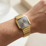 A gold-tone wristwatch with a square gray dial and matching gold-tone metal bracelet, shown worn on a person's wrist.