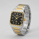 A men's two-tone stainless steel and gold watch with a black dial, featuring a square bezel, gold-tone hour markers and crown, and a matching two-tone metal link bracelet.