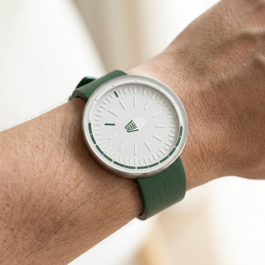 A minimalist wristwatch with a white dial, silver-toned case, and green strap, shown worn on a person's wrist.