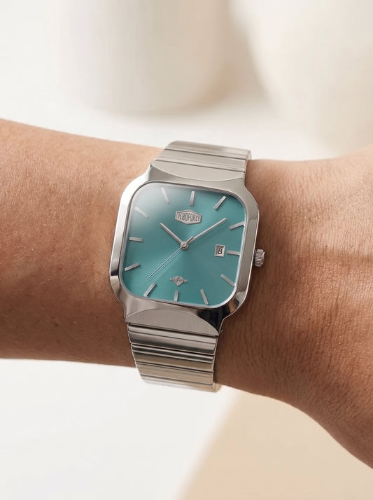 A silver-toned wristwatch with a turquoise dial and a matching silver link bracelet, shown worn on a person's wrist against a soft, light beige background.