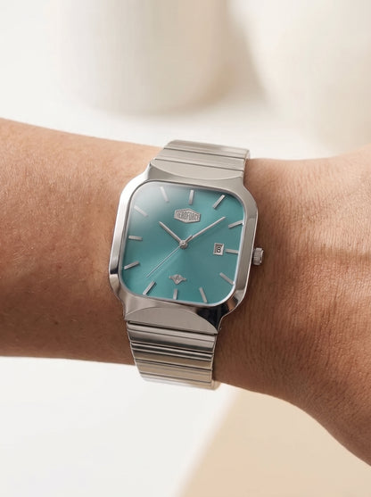 A silver-toned wristwatch with a turquoise dial and a matching silver link bracelet, shown worn on a person's wrist against a soft, light beige background.