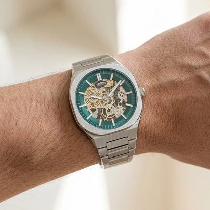 A silver-tone metal wristwatch with a green skeleton dial showing the inner mechanical movement, worn on a person's wrist.