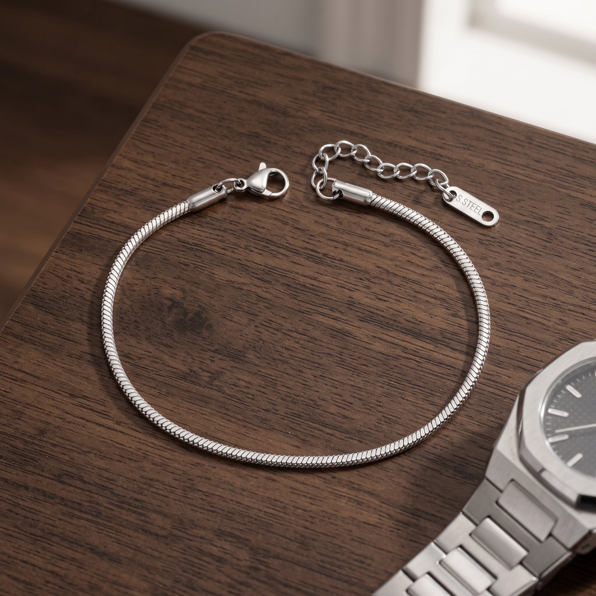 A silver-toned snake chain bracelet with a lobster clasp and extender chain, displayed on a dark wood surface next to a silver watch.
