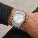 A silver-toned stainless steel wristwatch with an octagonal bezel and a white textured dial, shown worn on a person's wrist alongside a thin silver chain bracelet.