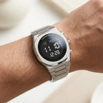 A silver stainless steel analog-digital wrist watch with a black round face, worn on a person's wrist against a soft, light beige background.