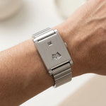 A silver stainless steel wristwatch with a rectangular case and integrated metal link bracelet, shown worn on a person's wrist. The watch features a small date window and a semi-circular index dial.