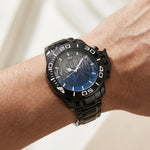 A wristwatch with a black bezel and black metal link bracelet, featuring a blue and black tiger-stripe pattern dial, shown worn on a person's wrist.