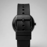 Close-up of a black watch strap buckle with the brand name debossed on the metal clasp, set against a soft gray background.