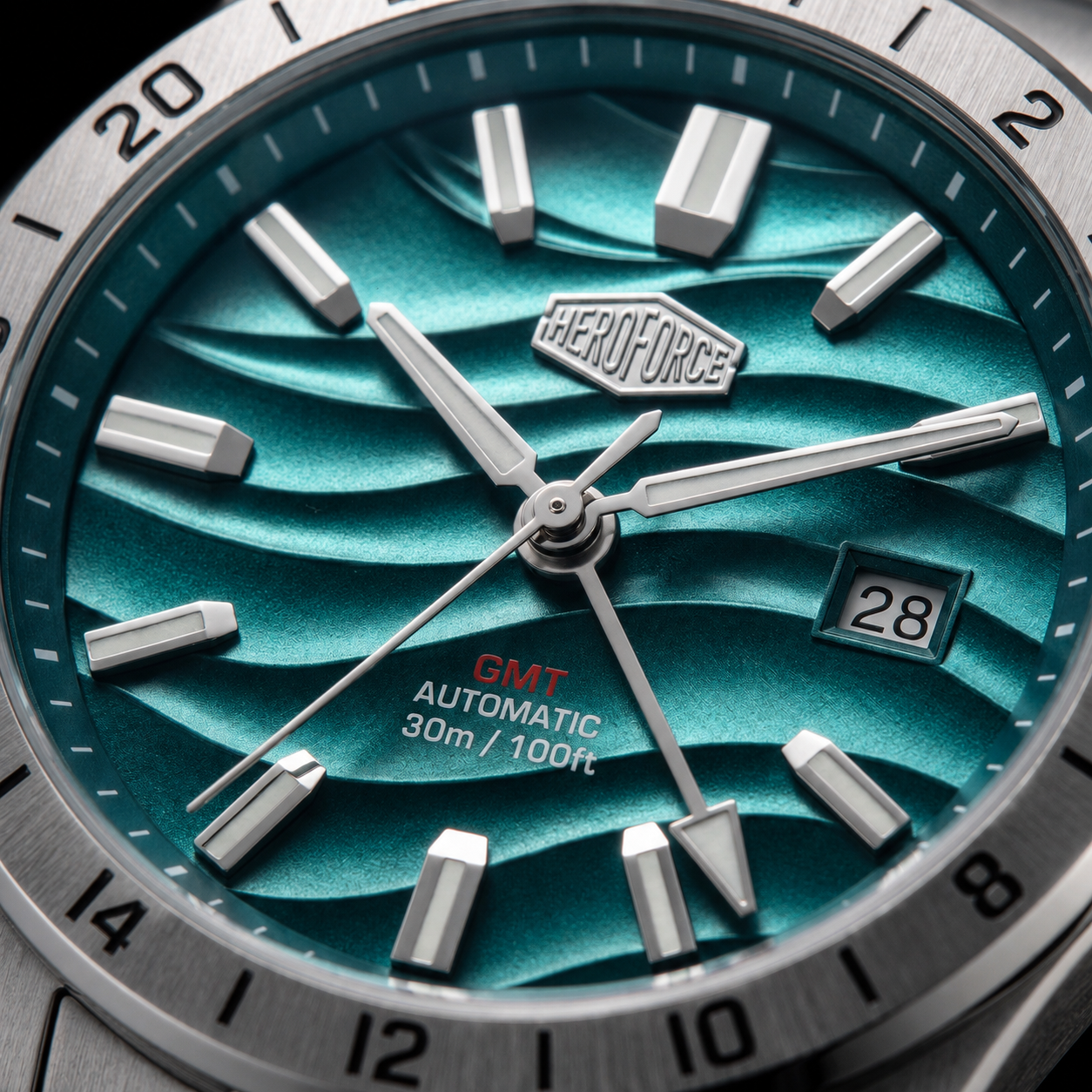 Close-up of a HeroForce GMT Automatic watch dial with a metallic teal wave-patterned face, silver baton hour markers, a date window, and a silver bezel.