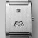 Close-up of a brushed stainless steel watch face with a small square day window showing 10 and a fan-shaped hour-index window below, on a light gray background.