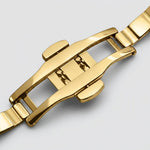 Close-up of a polished yellow gold stainless steel watch clasp and bracelet links on a light gray background.