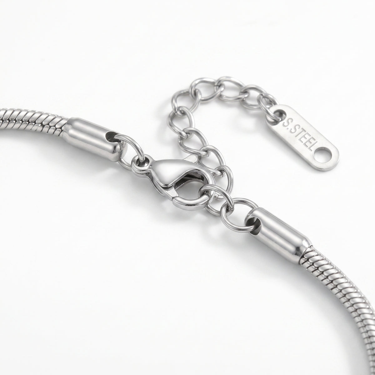 Close-up of a silver-toned snake chain bracelet showing the lobster clasp, extension chain, and a polished tag engraved with 'SS STEEL' .