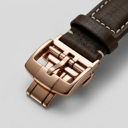 Close-up of a watch strap with a dark brown textured leather band and a polished rose gold buckle, shown on a light gray background.