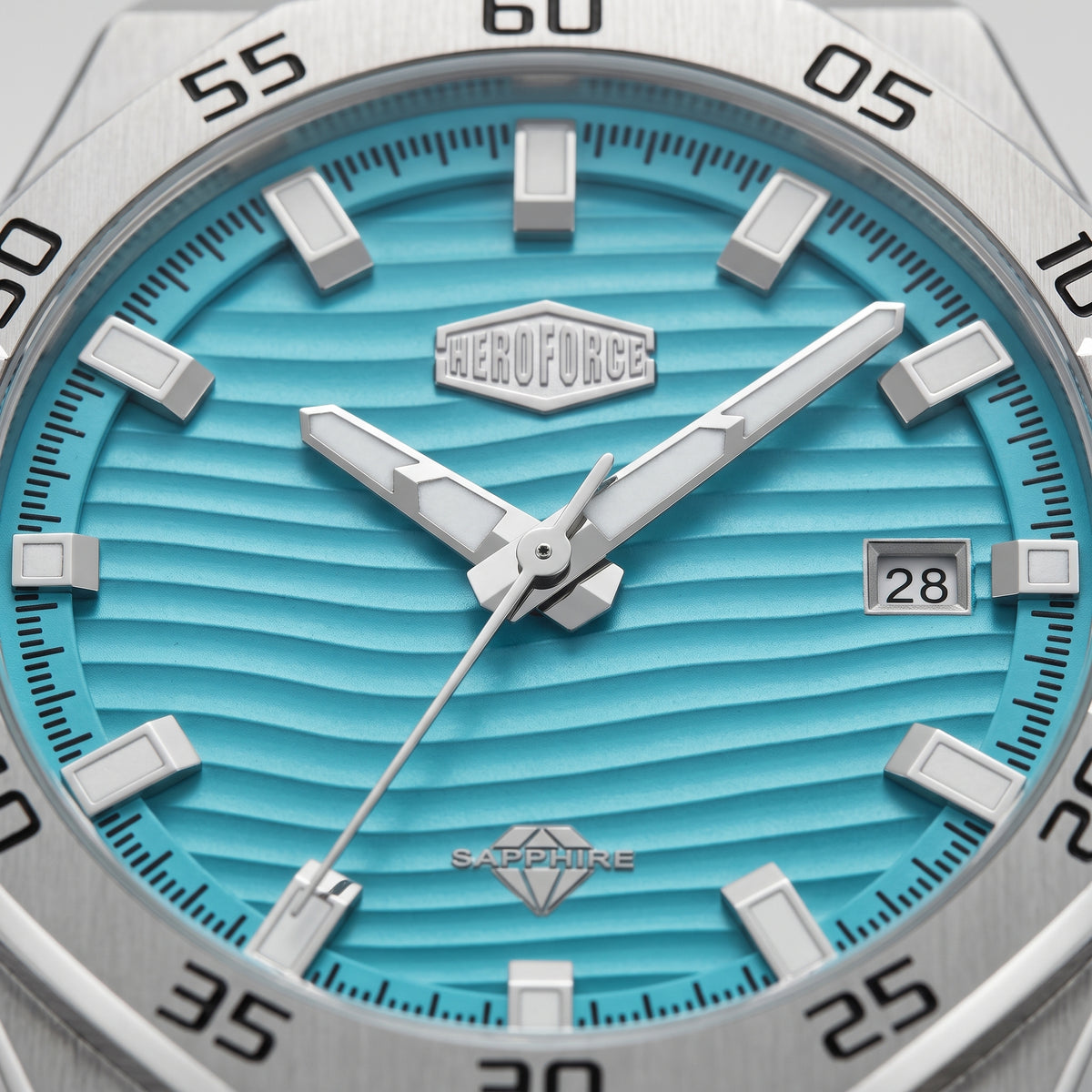 Close-up of a wristwatch dial with a bright turquoise blue wavy textured face, silver-tone hour markers and hands, a date window showing 28, and a stainless steel bezel with black minute markings.