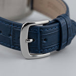 A close-up of a navy blue textured leather watch strap with a polished silver-tone metal buckle, resting on a neutral gray surface.