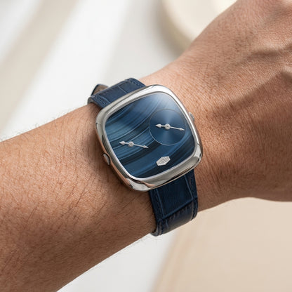 A close-up of a wrist wearing a modern square analog watch with a blue marbled dial, silver-toned case, and dark blue leather strap.