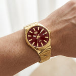A gold-tone wristwatch with a deep burgundy dial, shown worn on a person's wrist against a soft, light background.