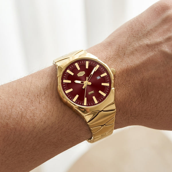 A gold-tone wristwatch with a deep burgundy dial, shown worn on a person's wrist against a soft, light background.