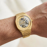 Wrist worn of HEROFORCE gold wristwatch with metallic bracelet, skeleton dial showcasing internal gears, gold markers and hands, date window at 6, elegant mechanical timepiece