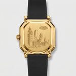 A gold-tone octagonal wristwatch shown from the back, featuring an engraved cityscape design
