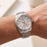 A silver stainless steel wristwatch with a textured white dial and orange hands, shown worn on a person's wrist.