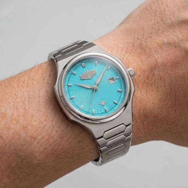 A silver stainless steel wristwatch with a textured turquoise blue dial, shown worn on a person's wrist against a neutral light gray background.