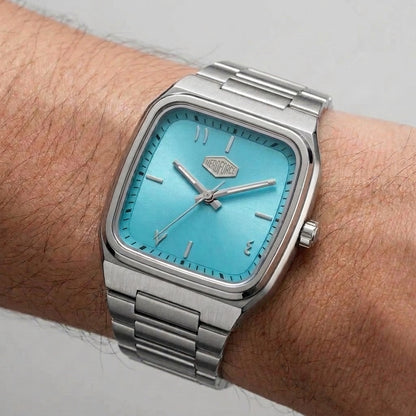 A silver stainless steel wristwatch with a bright turquoise blue dial, worn on a person's wrist against a neutral light gray background.