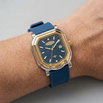 A wristwatch with a blue strap, blue dial, gold-tone bezel, and silver-tone case, shown worn on a person's wrist against a light gray background.