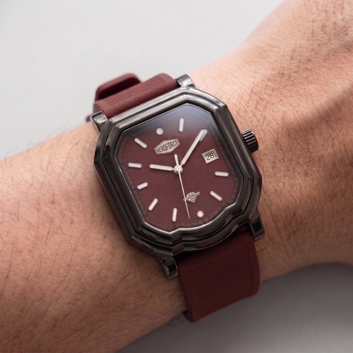 A wristwatch with a dark metallic octagonal case, a textured burgundy dial, and a matching burgundy strap, shown worn on a person's wrist against a light gray background.