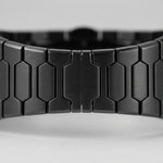 Close-up of a black metallic link watch band with a brushed finish and geometric hexagonal links, shown against a light gray background.