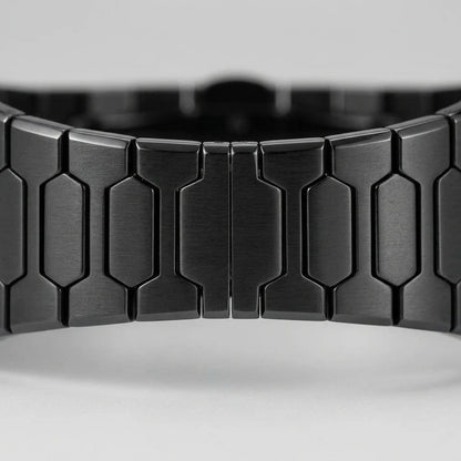 Close-up of a black metallic link watch band with a brushed finish and geometric hexagonal links, shown against a light gray background.