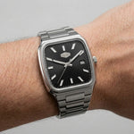 A silver-toned metal wristwatch with a black dial and rectangular cushion-shaped case, shown worn on a person's wrist