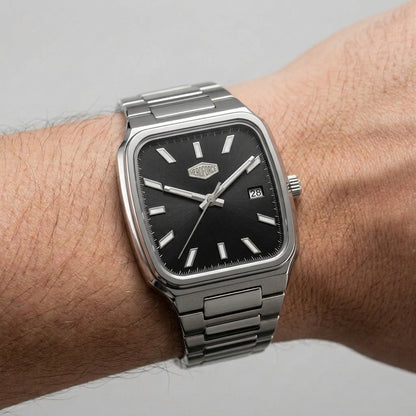 A silver-toned metal wristwatch with a black dial and rectangular cushion-shaped case, shown worn on a person's wrist