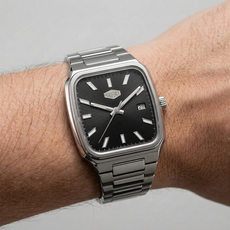 A silver-toned metal wristwatch with a black dial and rectangular cushion-shaped case, shown worn on a person's wrist