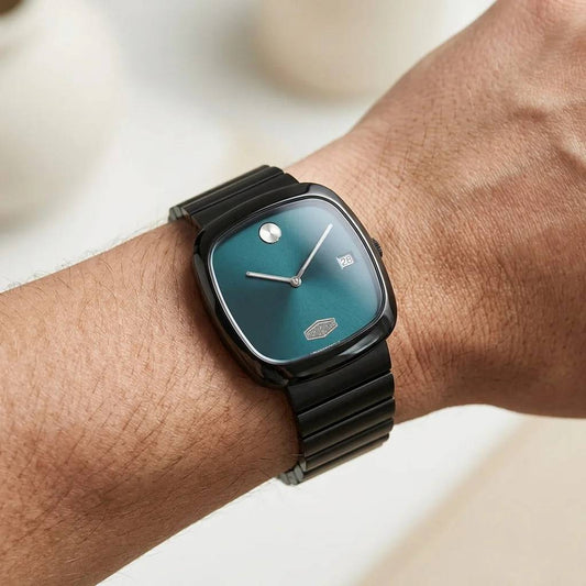 A close-up of a wrist wearing a Movado watch with a dark teal-green dial and a black metal link bracelet.