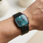 A close-up of a wrist wearing a Movado watch with a dark teal-green dial and a black metal link bracelet.